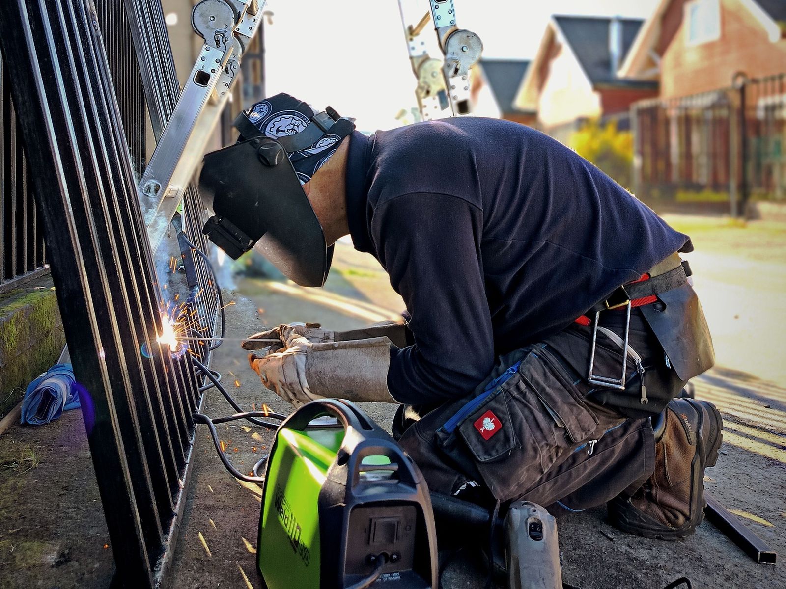 Welder in a helmet kneeling to repair a black iron fence panel on site, sparks at the joint, portable welder beside him
